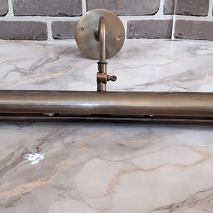 May include: A brass picture light fixture featuring a long, cylindrical horizontal shade attached to a circular wall-mounting plate by a single adjustable arm with a small thumb screw mechanism for positioning the angle of the light.