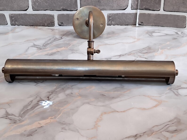 May include: A brass picture light fixture featuring a long, cylindrical horizontal shade attached to a circular wall-mounting plate by a single adjustable arm with a small thumb screw mechanism for positioning the angle of the light.