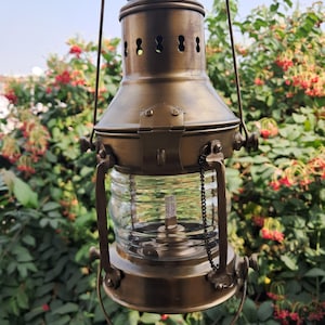 Peut inclure: Une lanterne en laiton de style vintage avec un globe en verre transparent et une poignée. La lanterne a un dessus décoratif avec des ouvertures d'aération et une petite chaîne. Le fond présente un feuillage vert et des fleurs rouges.