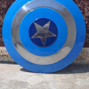 Op de afbeelding: Een rond schild in blauw, zilver en donkerblauw. Het schild heeft een zilveren ster in het midden. Het is een rekwisiet of verzamelobject, dat doet denken aan het iconische accessoire van een superheld. De diameter is ongeveer 76 cm.
