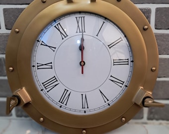 May include: A round, gold-colored clock resembling a ship's porthole. The clock face is white with black Roman numerals and hands. The clock is mounted on a gray brick wall.