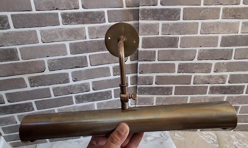 May include: A brass picture light fixture featuring a cylindrical horizontal shade attached to a vertical arm with an adjustable knuckle joint and a circular wall-mounting plate. The metal has a muted, brushed finish with a warm golden-brown tone.