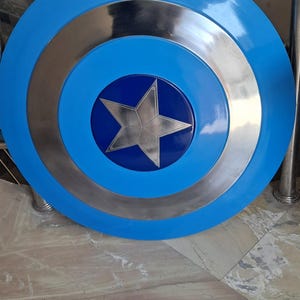 Op de afbeelding: Een rond, blauw en zilver schild met een grote zilveren ster in het midden. Het schild heeft concentrische cirkels van blauw en zilver. Een klassiek superheldenaccessoire.