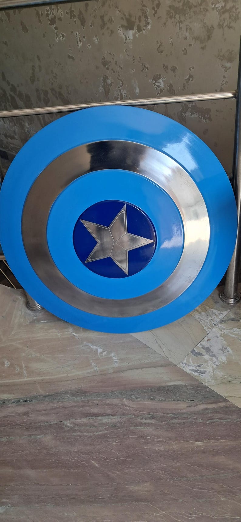 Op de afbeelding: Een rond, blauw en zilver schild met een grote zilveren ster in het midden. Het schild heeft concentrische cirkels van blauw en zilver. Een klassiek superheldenaccessoire.