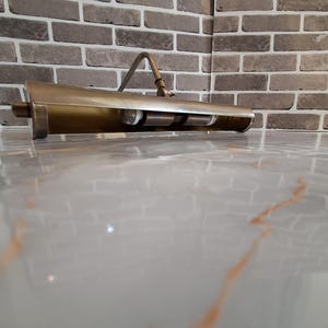 May include: A brass-toned metal picture light with a long, cylindrical shade and an adjustable arm. The fixture features a metallic finish and contains two internal light bulb sockets visible within the open underside of the horizontal housing.