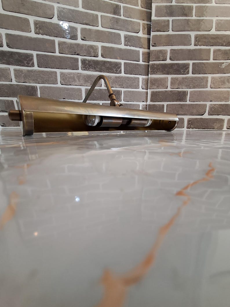 May include: A brass-toned metal picture light with a long, cylindrical shade and an adjustable arm. The fixture features a metallic finish and contains two internal light bulb sockets visible within the open underside of the horizontal housing.