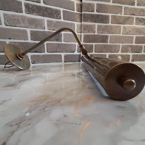 May include: A brass-toned wall-mounted picture light featuring a cylindrical lamp head and an adjustable arm. The fixture includes a circular mounting plate with exposed wiring and a cylindrical shade with a knob on the end, finished in a metallic tone.