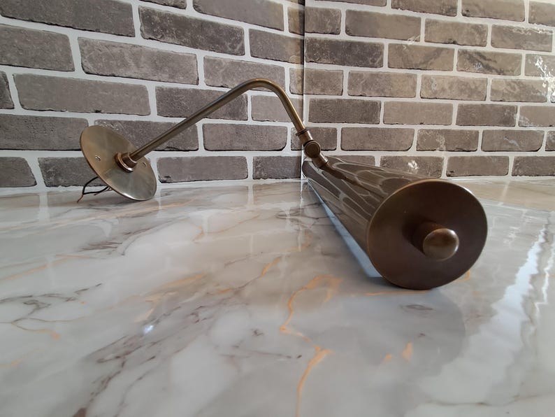 May include: A brass-toned wall-mounted picture light featuring a cylindrical lamp head and an adjustable arm. The fixture includes a circular mounting plate with exposed wiring and a cylindrical shade with a knob on the end, finished in a metallic tone.