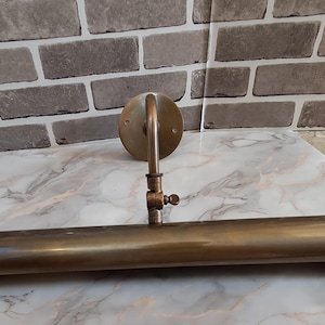 May include: A wall-mounted picture light with a cylindrical brass-toned metal shade and a circular mounting plate. The fixture features an adjustable arm with a central hinge and a small screw knob for positioning the light angle precisely.