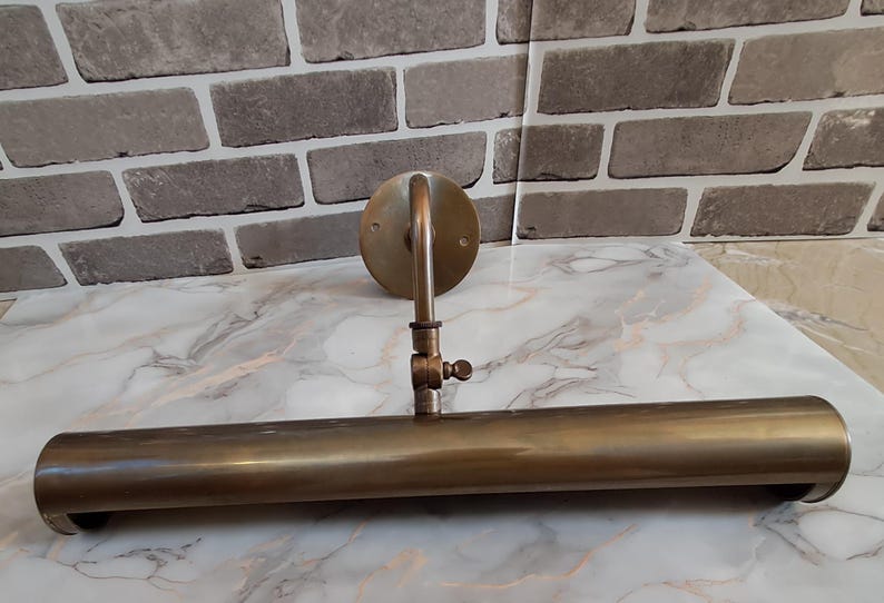 May include: A wall-mounted picture light with a cylindrical brass-toned metal shade and a circular mounting plate. The fixture features an adjustable arm with a central hinge and a small screw knob for positioning the light angle precisely.