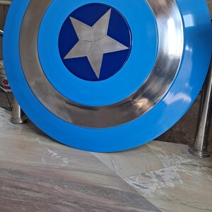 Op de afbeelding: Een replica van het Captain America-schild. Het schild is rond, met een helderblauwe buitenring, een zilveren binnenring en een centrale blauwe cirkel met een zilveren ster. Het schild ligt op een marmeren oppervlak.