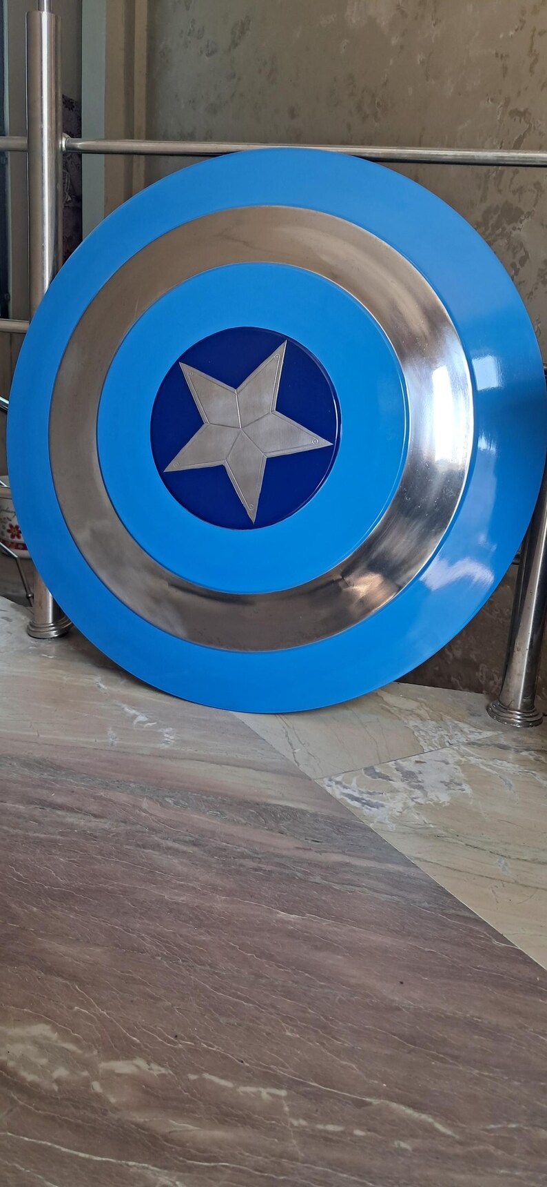 Op de afbeelding: Een replica van het Captain America-schild. Het schild is rond, met een helderblauwe buitenring, een zilveren binnenring en een centrale blauwe cirkel met een zilveren ster. Het schild ligt op een marmeren oppervlak.