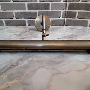May include: A wall-mounted picture light fixture with a cylindrical, antique brass-toned metal shade. The lamp features a circular mounting plate, a curved support arm, and an adjustable hinge mechanism with a small screw for positioning the light.