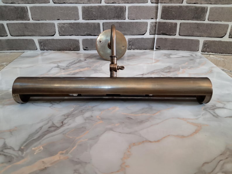 May include: A wall-mounted picture light fixture with a cylindrical, antique brass-toned metal shade. The lamp features a circular mounting plate, a curved support arm, and an adjustable hinge mechanism with a small screw for positioning the light.