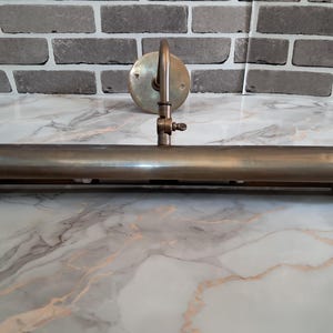 May include: A brass picture light fixture featuring a long, cylindrical horizontal shade attached to a circular wall-mounting plate by a single adjustable arm with a small wing nut screw for positioning the angle of the light.