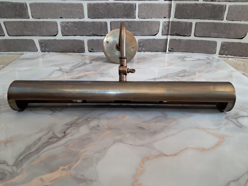 May include: A brass picture light fixture featuring a long, cylindrical horizontal shade attached to a circular wall-mounting plate by a single adjustable arm with a small wing nut screw for positioning the angle of the light.