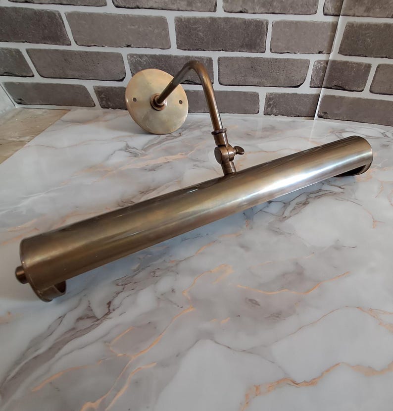 May include: A brass picture light with a cylindrical shade and an adjustable arm. The fixture features a circular wall-mounting plate and a central hinge mechanism that allows for tilting the light tube to direct illumination as needed.