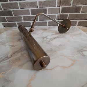 May include: A metallic bronze picture light with a cylindrical lamp head, an adjustable arm, and a circular wall mounting plate. The fixture features a smooth, polished finish and a small knob on the end of the tube for manual operation.