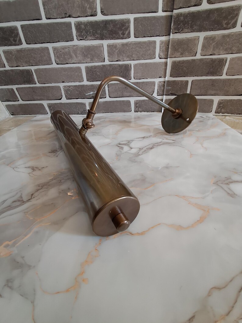 May include: A metallic bronze picture light with a cylindrical lamp head, an adjustable arm, and a circular wall mounting plate. The fixture features a smooth, polished finish and a small knob on the end of the tube for manual operation.