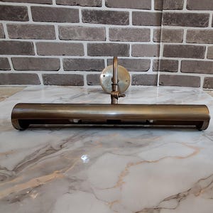 May include: A brass picture light fixture featuring a long, cylindrical horizontal shade attached to a circular wall-mounting plate by an adjustable arm. The metal has a muted, aged finish, and the design includes a visible hinge mechanism.