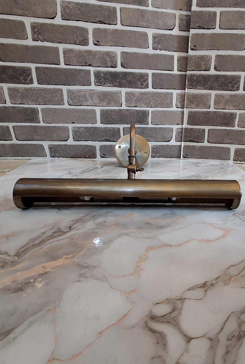 May include: A brass picture light fixture featuring a long, cylindrical horizontal shade attached to a circular wall-mounting plate by an adjustable arm. The metal has a muted, aged finish, and the design includes a visible hinge mechanism.