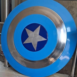Op de afbeelding: Een rond, blauw schild met zilveren en blauwe concentrische cirkels. Een zilveren vijfpuntige ster is gecentreerd op een donkerblauwe cirkel. Het schild heeft een glanzende afwerking en is waarschijnlijk een rekwisiet of verzamelobject.