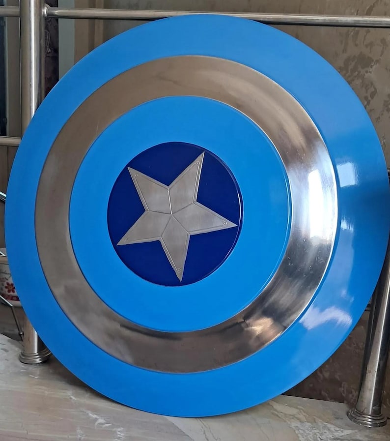 Op de afbeelding: Een rond, blauw schild met zilveren en blauwe concentrische cirkels. Een zilveren vijfpuntige ster is gecentreerd op een donkerblauwe cirkel. Het schild heeft een glanzende afwerking en is waarschijnlijk een rekwisiet of verzamelobject.