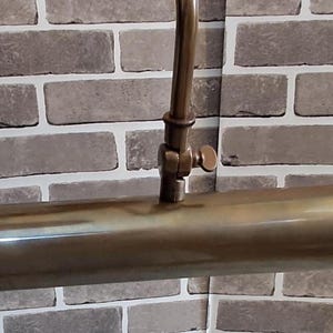 May include: A tubular picture light with a metallic brass finish. The fixture features a long, cylindrical lamp head attached to a central adjustable arm with a small screw knob, allowing for precise positioning of the light source.