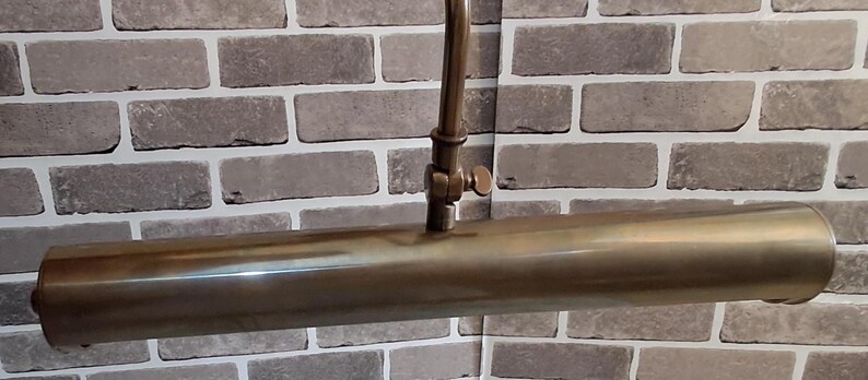 May include: A tubular picture light with a metallic brass finish. The fixture features a long, cylindrical lamp head attached to a central adjustable arm with a small screw knob, allowing for precise positioning of the light source.