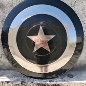 Puede incluir: Un escudo redondo negro y plateado con una estrella plateada central. El escudo tiene un borde exterior negro, un anillo plateado y un centro negro con una estrella plateada. Descansa sobre una superficie texturizada.