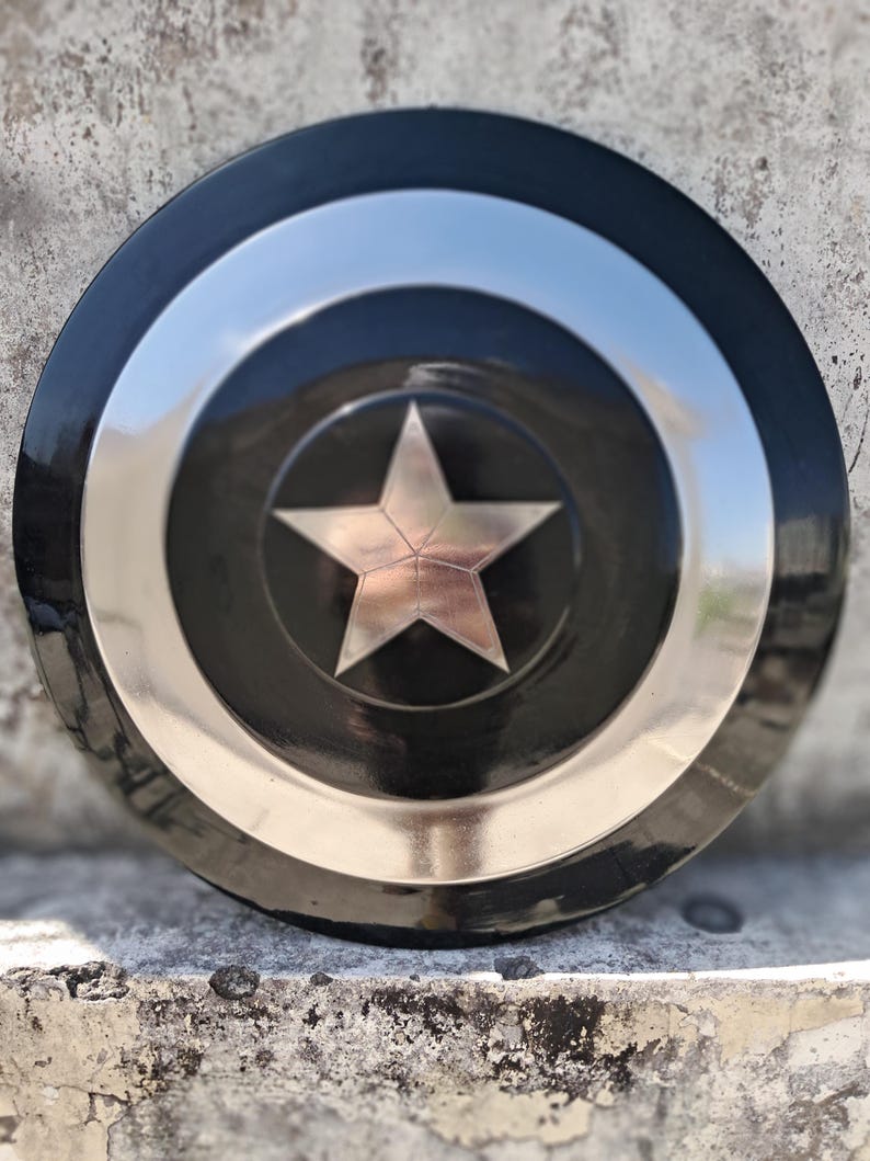 Puede incluir: Un escudo redondo negro y plateado con una estrella plateada central. El escudo tiene un borde exterior negro, un anillo plateado y un centro negro con una estrella plateada. Descansa sobre una superficie texturizada.