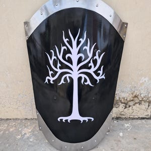 Pode incluir: Um escudo preto com um desenho de árvore branca. O escudo tem uma parte superior e inferior curvas com uma moldura e rebites de metal prateado. O desenho da árvore está centrado.