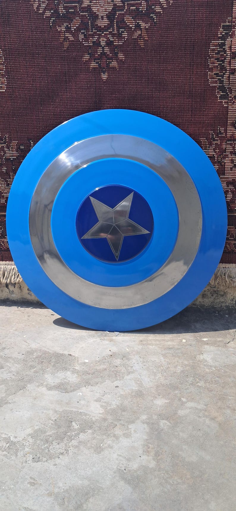 Op de afbeelding: Een rond schild in blauw, zilver en donkerblauw. Het schild heeft een zilveren ster in het midden. Dit rekwisiet of verzamelobject doet denken aan het iconische accessoire van een superheld.