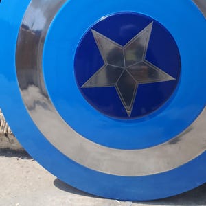 Op de afbeelding: Een blauw en zilver schild met een centraal sterontwerp. Het schild heeft concentrische cirkels van blauw en zilver, met een donkerblauw centrum met een zilveren ster. Een klassiek superheldenaccessoire.