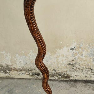 Pode incluir: Um bastão de madeira artesanal esculpido em forma de cobra. A cabeça da cobra é detalhada com escamas e olhos esculpidos. O corpo da cobra curva-se graciosamente, com padrões de escamas detalhados. A madeira tem um tom castanho quente.