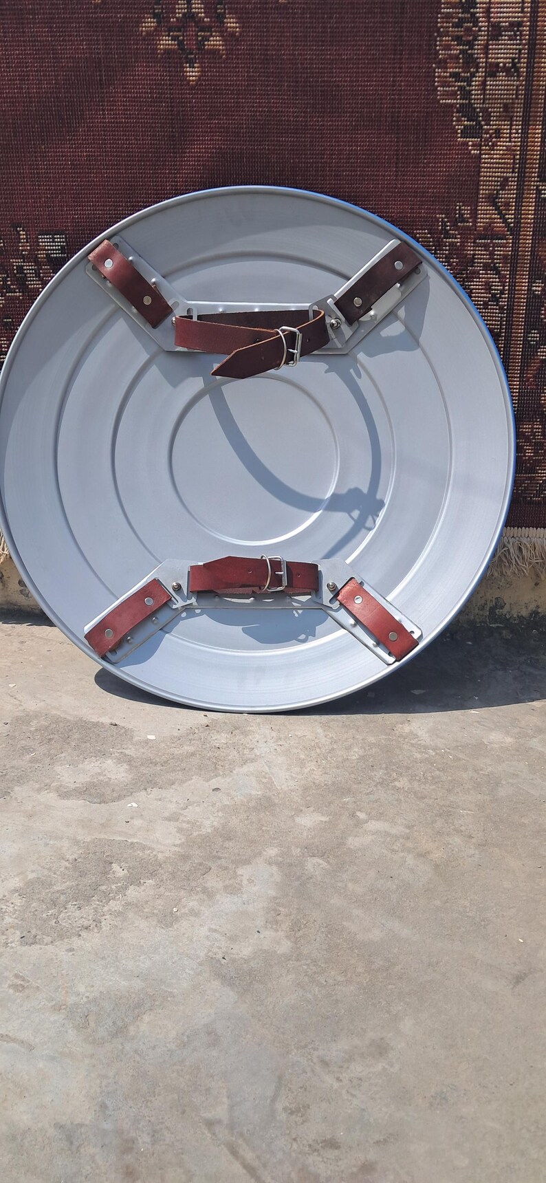 Op de afbeelding: Een zilverkleurig, rond schild met concentrische cirkels. Het schild heeft vier bruine leren riemen met zilveren gespen en klinknagels om het vast te zetten. Het schild is waarschijnlijk van metaal. Diameter ongeveer 76 cm.