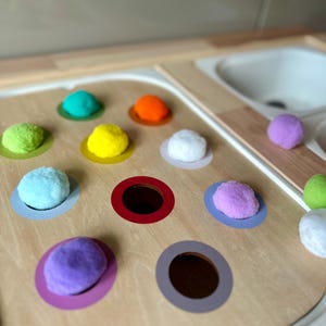 Könnte beinhalten: Ein Holzspielzeug mit bunten Pompons und passenden farbigen Kreisen. Die Pompons sind in den Farbtönen Grün, Blau, Gelb, Orange, Weiß und Lila gehalten. Das Spielzeug hat Löcher für die Pompons und befindet sich in der Nähe eines weißen Waschbeckens.