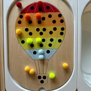 Könnte beinhalten: Ein Holzpuzzle mit einem bunten Heißluftballon-Design. Der Ballon ist in Abschnitte aus Rot, Orange, Gelb und Blau unterteilt. Schwarze Punkte befinden sich auf dem Ballon. Bunte Pompons sind um das Puzzle herum verstreut.