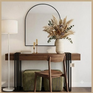 May include: A wooden console table with black metal legs, an arched wall mirror, a floor lamp with a white shade, a wooden chair, two olive green corduroy ottomans, and a textured ceramic vase filled with dried pampas grass and foliage.