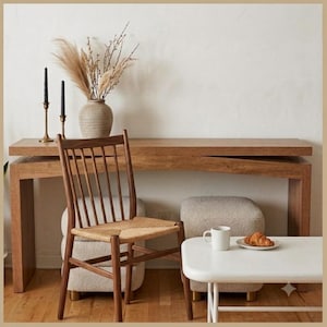 May include: A wooden console table with a natural finish, a spindle-back chair with a woven seat, two beige textured ottomans, and a white coffee table. A ceramic vase with dried grasses, two black candles, and a croissant on a plate are present.