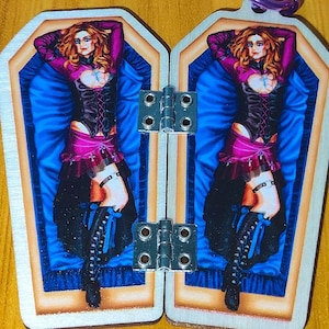 Peut inclure: Deux pendentifs en forme de cercueil articulés avec une bordure argentée. Chaque pendentif représente une femme en corset et bottes, allongée sur une surface bleue satinée. La femme a les cheveux roux et porte une tenue noire et rose.