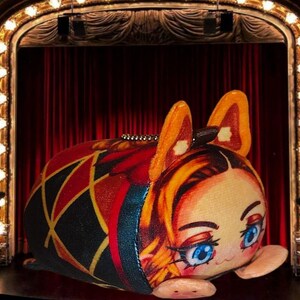 Peut inclure: Une peluche avec un visage de dessin animé et de grands yeux bleus. Le jouet a des oreilles et des cheveux orange, et un corps à motifs avec des formes de diamants rouges, noirs et jaunes. Une chaîne argentée est attachée au sommet. Le fond présente une scène avec des rideaux rouges.