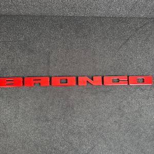 Puede incluir: Emblema "BRONCO" de color rojo brillante. Las letras son piezas individuales con un acabado brillante. El emblema se muestra sobre una superficie gris oscuro y texturizada.