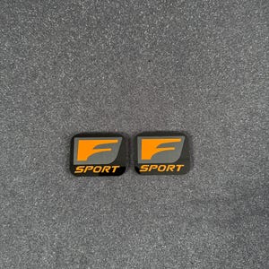 Könnte beinhalten: Zwei schwarze und orangefarbene "Sport"-Embleme. Jedes Emblem hat einen schwarzen Hintergrund mit dem Wort "SPORT" in weißem Text unten. Die Oberseite jedes Emblems hat ein orangefarbenes "F"-Design.