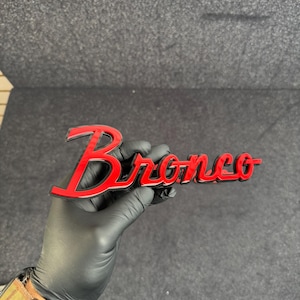 May include: A red and black "Bronco" emblem, held by a gloved hand. The emblem features a glossy red finish with a black outline, showcasing the word "Bronco" in a stylized script. The background is a dark gray surface.