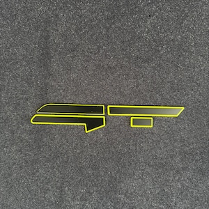 Puede incluir: Piezas de emblema de coche negras y amarillas. Las piezas son negras con un contorno amarillo. El emblema está en tres piezas separadas. El fondo es una superficie texturizada de color gris oscuro.