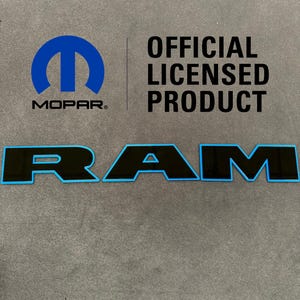 Może przedstawiać: Czarny emblemat "RAM" z jasnoniebieskim obrysem i logo Mopar. Widoczny jest również tekst "OFFICIAL LICENSED PRODUCT". Emblemat znajduje się na szarej powierzchni.