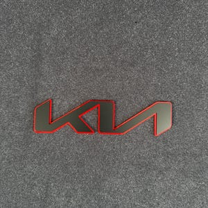 Puede incluir: Un emblema Kia negro y rojo. El emblema presenta el logotipo de Kia en un diseño estilizado con un contorno negro y un interior rojo. El emblema está sobre un fondo gris.