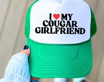 Peut inclure: Casquette de camionneur verte et blanche avec le texte "I ❤️ MY COUGAR GIRLFRIEND" en noir et rouge. La casquette a une visière et un dos en filet verts, avec un panneau avant blanc.