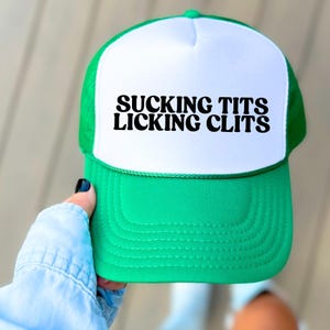 May include: A green and white trucker hat with the text "SUCKING TITS LICKING CLITS" in bold black letters. The hat has a white front panel and a green mesh back and brim.